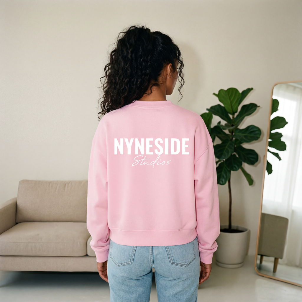 Person wearing a pink sweatshirt with 'Nyneside Studios' text, standing in a room with a couch and plant. Essentials women’s crew neck jumper, back view, relaxed fit knitwear
